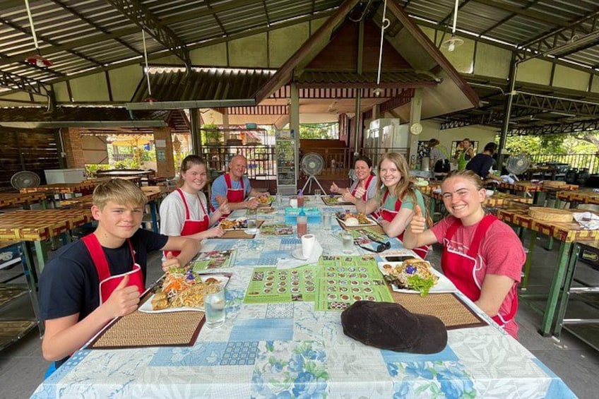 A group of people sitting at a long table with food and drinks, in an outdoor setting with a metal roof and wooden benches. 4