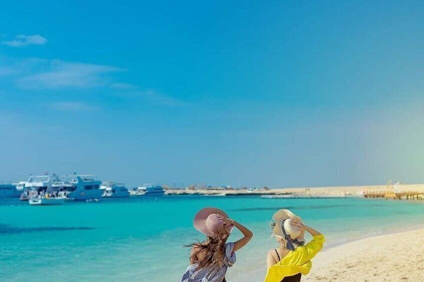 A beach with clear turquoise water, a sandy shore, and yachts in the background. 1