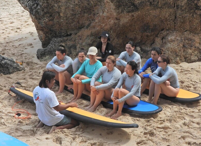 A group of people sitting on surfboards on a sandy beach with rocks in the background. 2
