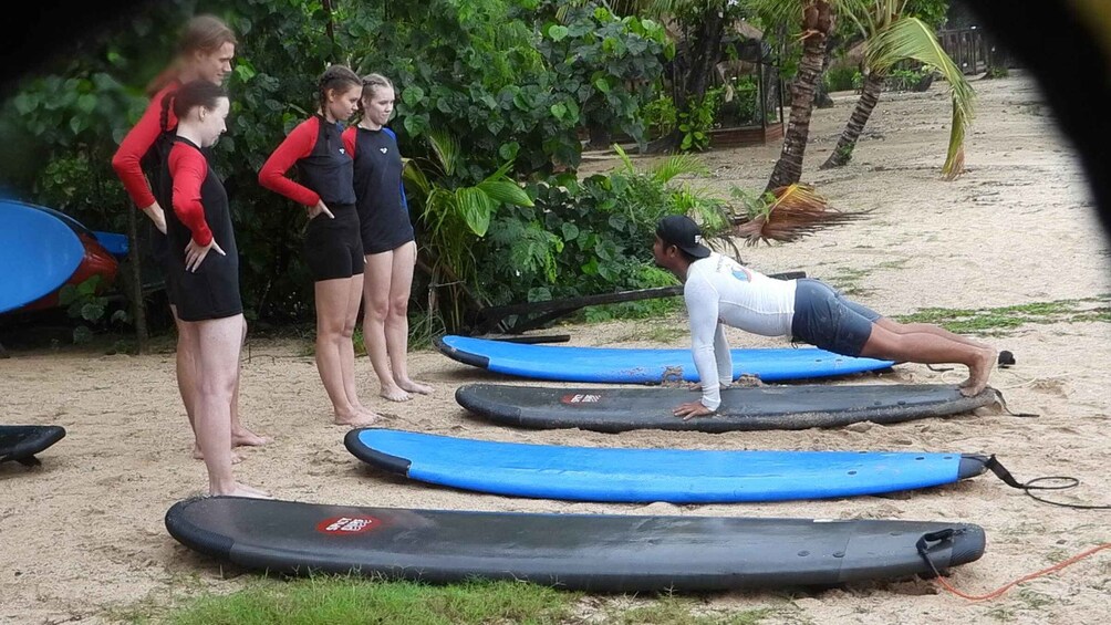 A person doing a push-up on a surfboard, surrounded by other people and surfboards on a sandy beach with greenery in the background. 4