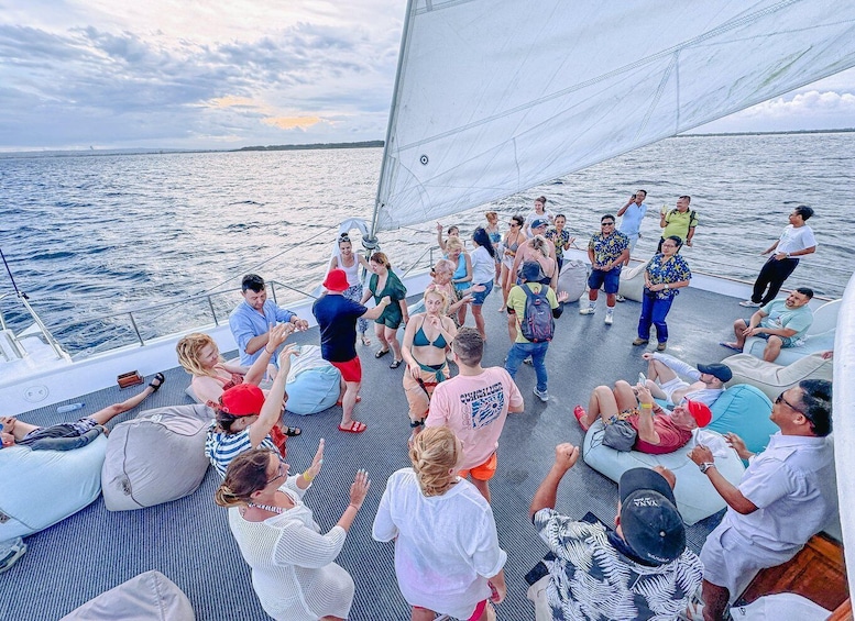 A group of people on a sailboat, some standing and others sitting, with the ocean and a cloudy sky in the background. 2