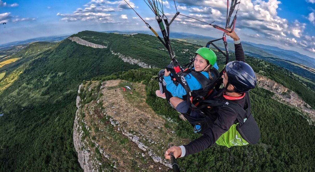 Two people paragliding over a mountainous landscape with green valleys and rocky outcrops. 1