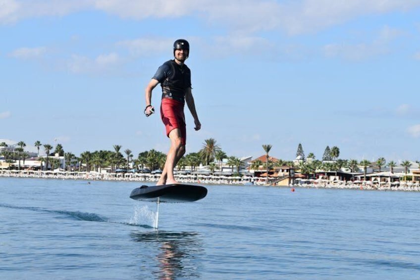 En person som åker wakeboard på ett vattendrag med en strand och byggnader i bakgrunden. 1