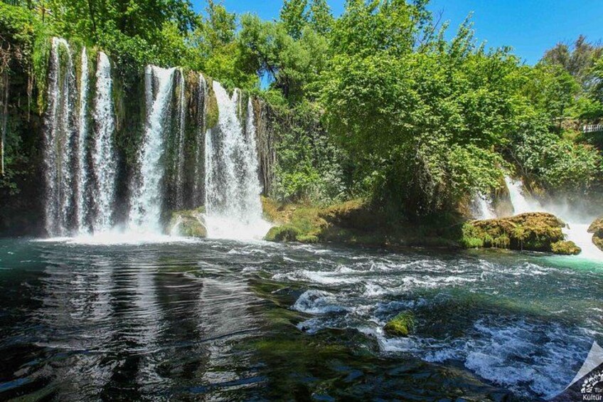 Ein Wasserfall stürzt über Felsen in einen Fluss, umgeben von üppiger Vegetation. 3