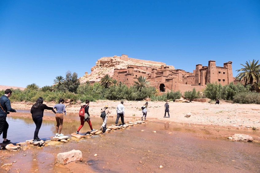 A group of people crossing a shallow water body in a desert landscape with a large, ancient fort in the background. 3