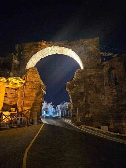 An ancient stone archway with a paved road leading through it, surrounded by stone walls and a lit building. 3