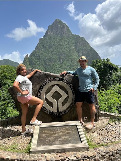 Two people posing in front of a large rock with a circular emblem and a plaque, with a mountain in the background. 5
