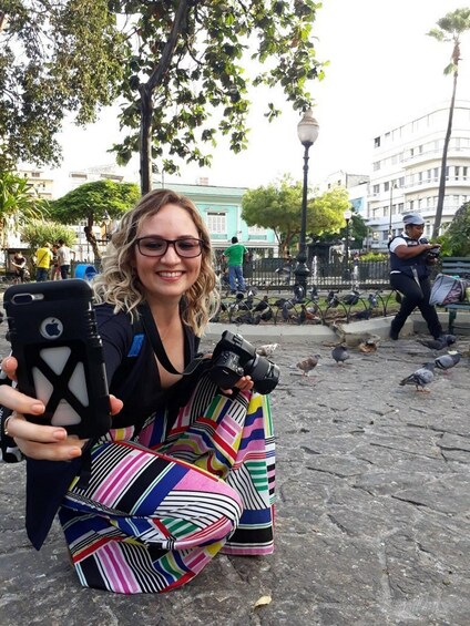 A person sitting on a stone surface, holding a smartphone and a camera. 1