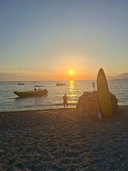 A sunset over a beach with a surfboard leaning against a rock and a person standing in the water. 2