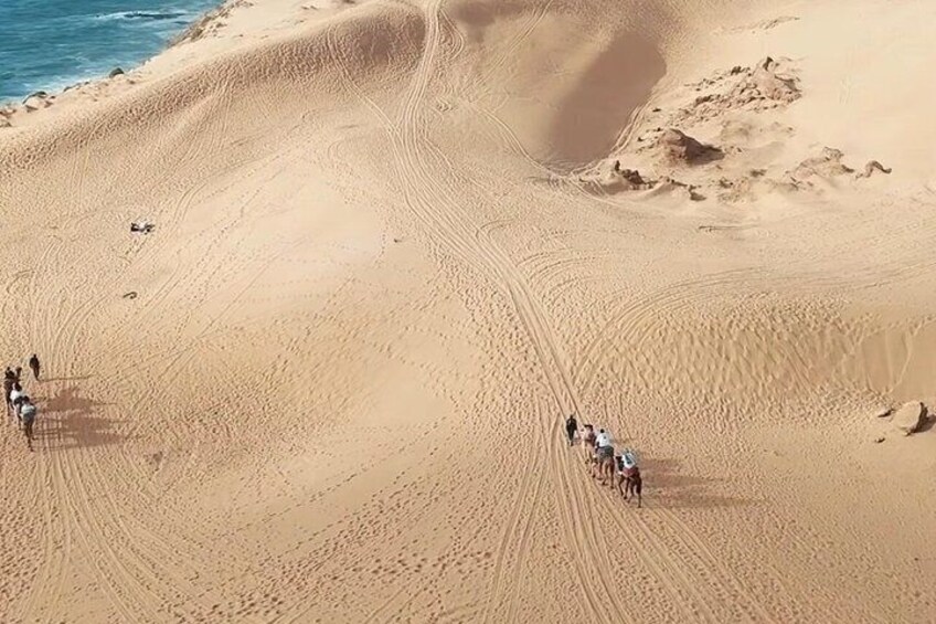 Un groupe de personnes à cheval traverse un paysage désertique sablonneux, avec des dunes ondulantes et une étendue d’eau en arrière-plan. 4