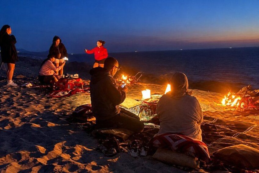 Des personnes sont assises sur une plage au coucher du soleil, avec un feu de camp en premier plan. 3