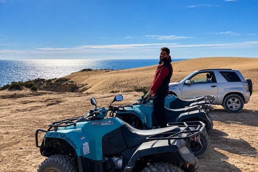 En person står bredvid en turkos fyrhjuling och en vit SUV på en sandig yta, med havet i bakgrunden. 5