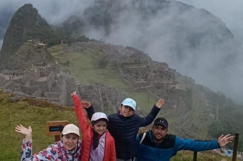 En gruppe mennesker står foran Machu Picchu med armene i vejret, omgivet af tåge og gamle ruiner. 2