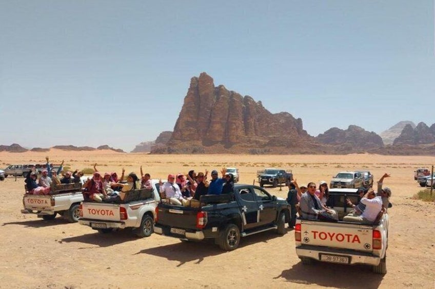 Woestijnlandschap met een opvallende rotsformatie op de achtergrond en verschillende Toyota pick-up trucks met passagiers. 3