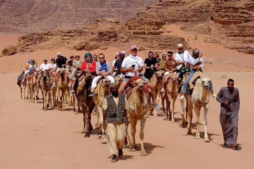 Een groep mensen die op kamelen door een woestijnlandschap rijdt, met rotsachtig terrein. 1