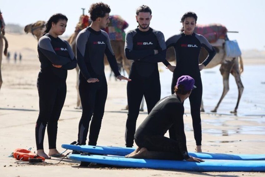 Personas con trajes de neopreno en una playa, con tablas de surf y un camello al fondo. 4