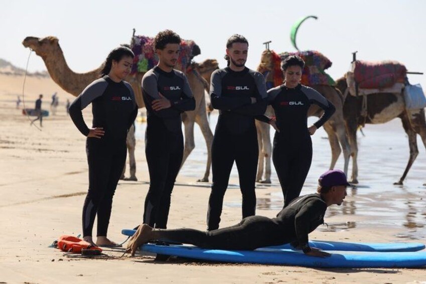 Escena de playa con gente en trajes de neopreno, una tabla de surf azul y camellos al fondo. 3