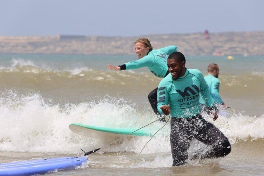 Personas surfeando en la playa, con olas y un paisaje urbano al fondo. 1