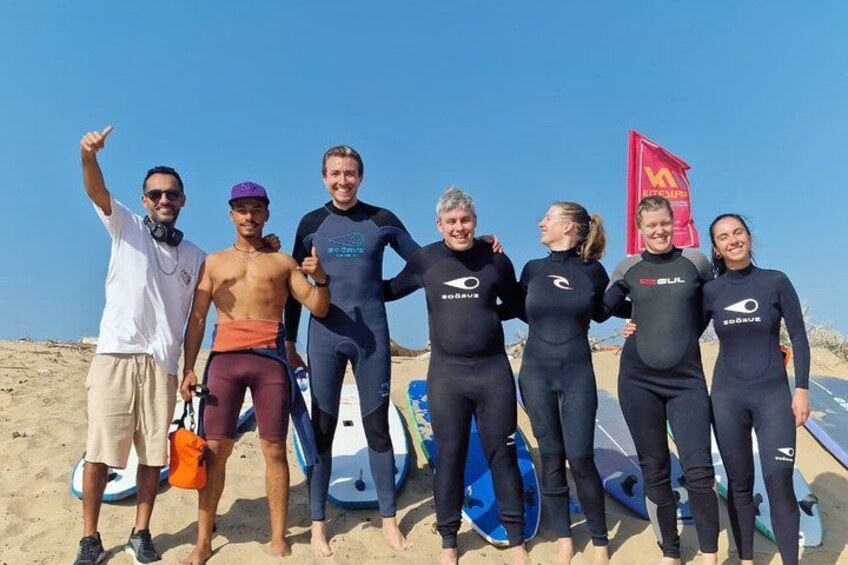 Un grupo de personas con trajes de neopreno en una playa, con tablas de surf y una bandera roja al fondo. 5