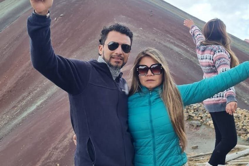 A person in a dark jacket taking a selfie with a woman in a teal jacket, and another person in a patterned sweater in the background. 3