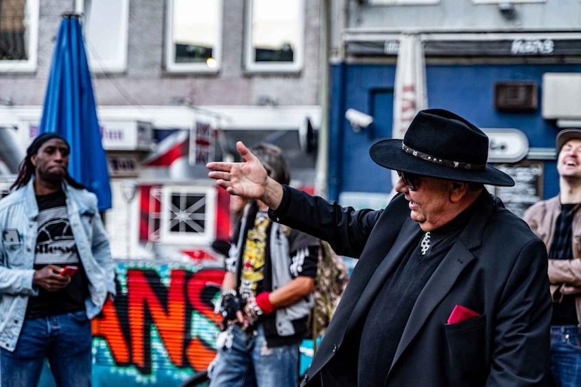 A person in a black hat and suit gesturing with their hand. 1