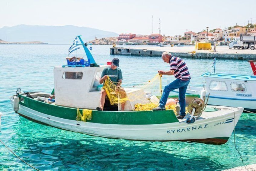 Un hombre pesca en un bote llamado 'KYKAONNAZ 1047' en un puerto con aguas azules cristalinas y un pueblo costero al fondo. 2