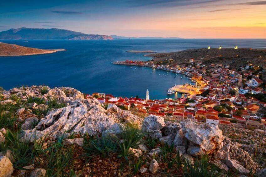 Un pueblo costero con edificios de techo rojo, un primer plano rocoso y vistas al mar y montañas lejanas. 3