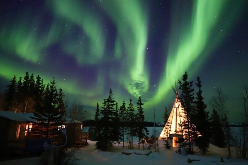 A snow-covered landscape with a wooden cabin and a teepee illuminated by warm lights, under a night sky with the aurora borealis. 2