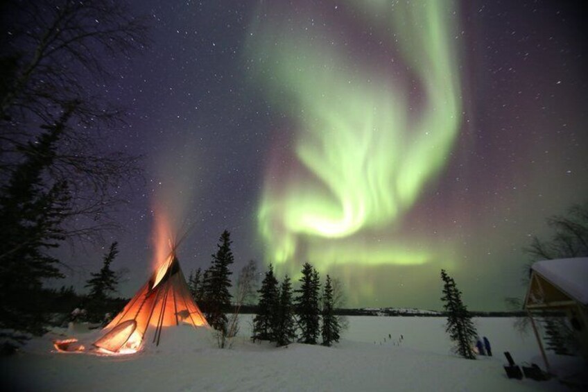 A snow-covered landscape with a lit campfire and a teepee, under a night sky with the Northern Lights. 1