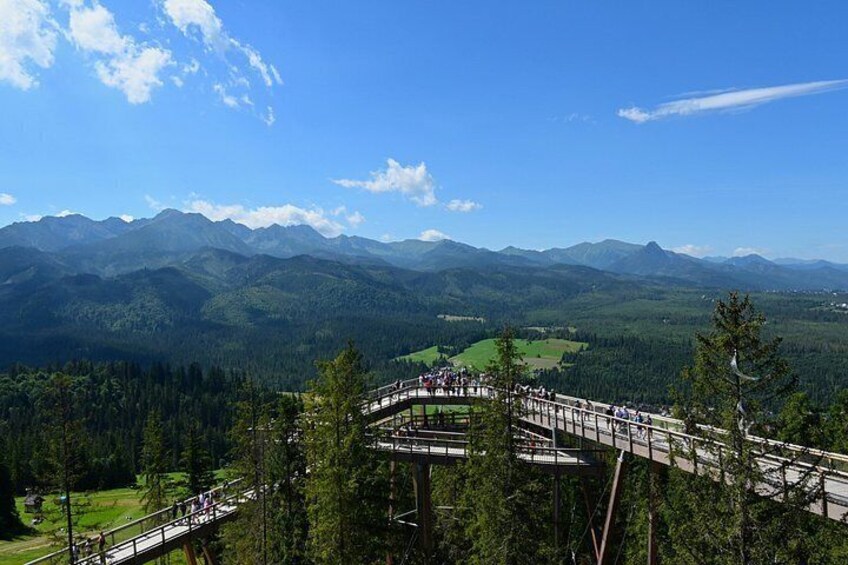 Eine Holzbrücke, über die Menschen gehen, umgeben von Bergen und Grün. 5
