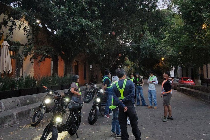 A group of people standing around parked motorcycles on a tree-lined street. 3