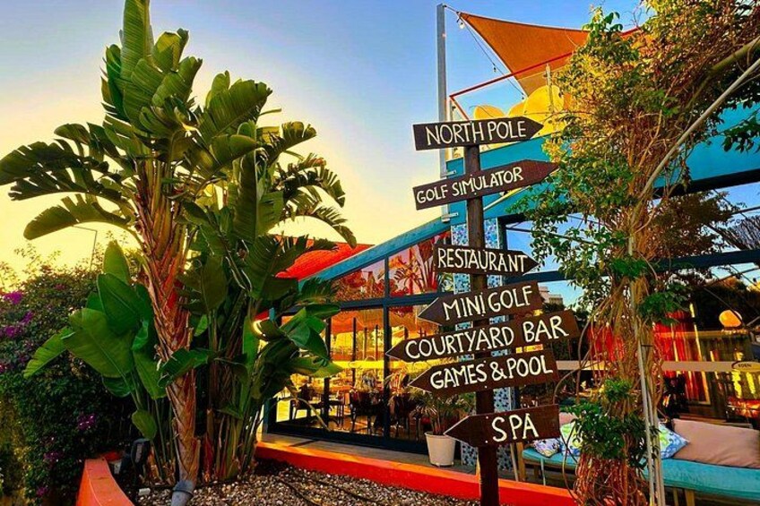 Un cartel con indicaciones hacia varios servicios, incluyendo simulador de golf, restaurante, mini golf, bar en el patio, juegos y piscina, y spa. 2