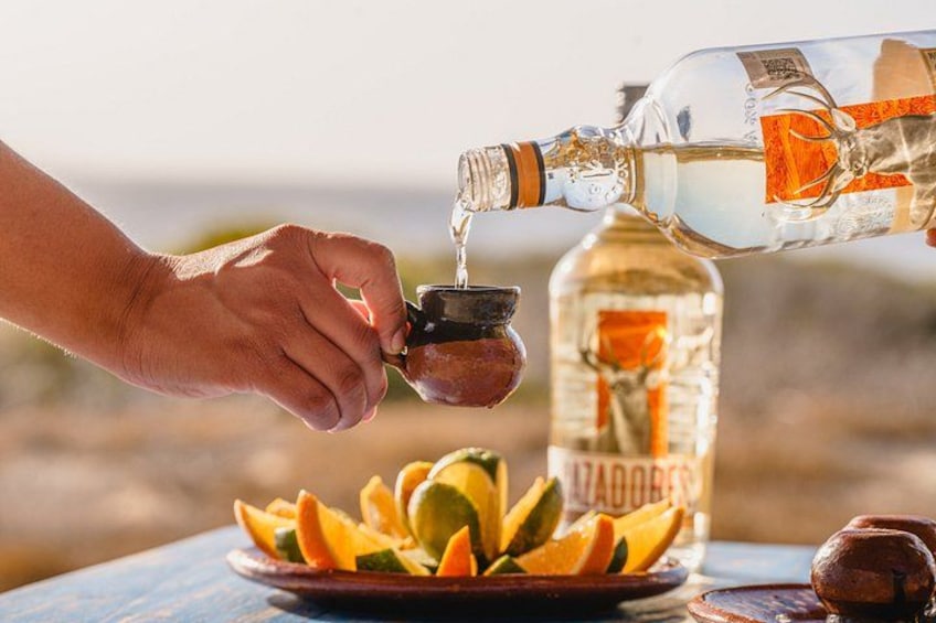 A hand pouring a clear liquid from a bottle into a small container. In the background, there is a bottle labeled 'AZADOGER' and a plate with sliced fruit. 2