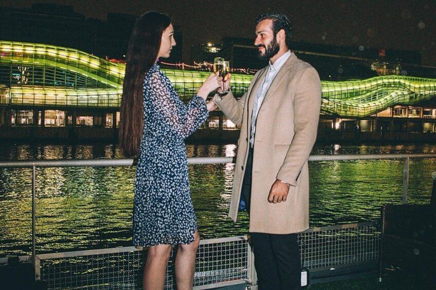 Une personne prend une photo avec un appareil photo, une autre personne tient une boisson, et un pont illuminé se trouve à l’arrière-plan. 1
