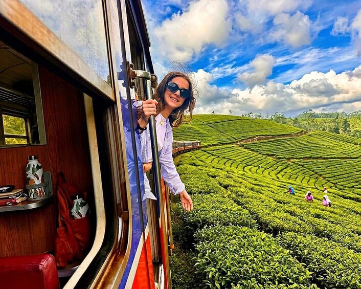 A person leaning out of a train window, holding onto the window frame, with a view of tea plantations and a blue sky with clouds. 1