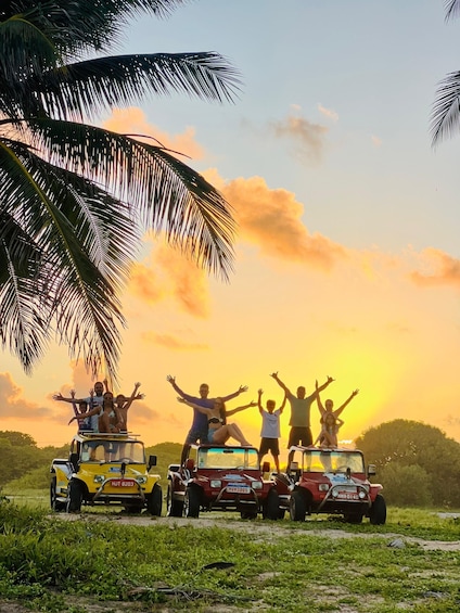A group of people on a vehicle with raised arms, palm trees, and a sunset. 2