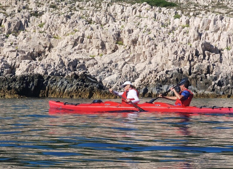 Two people kayaking in a red kayak on calm water with a rocky shoreline in the background. 2