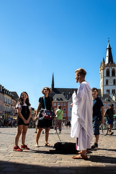 A group of people in a cobblestone square with historic buildings and a church tower in the background. 5