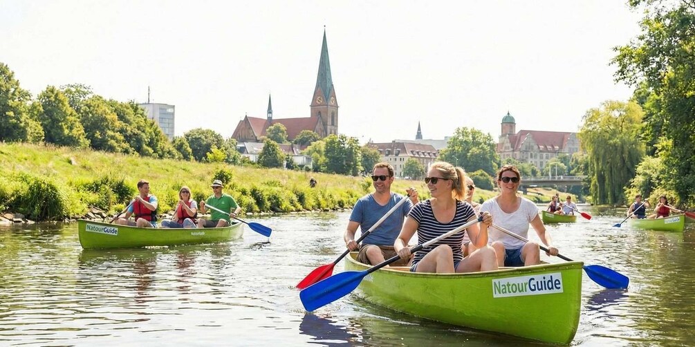 People in green canoes on a river with a church and buildings in the background. 1