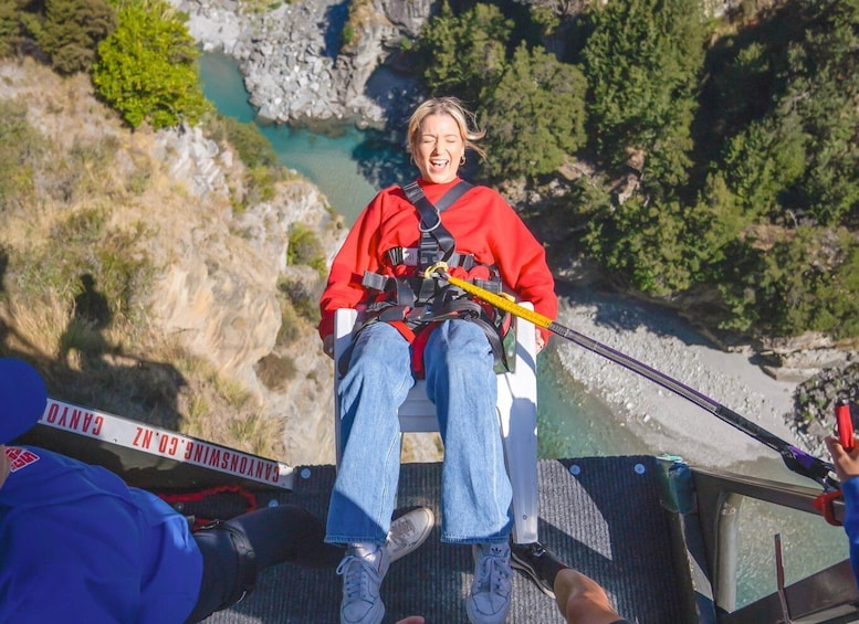 A person in a red jacket and blue jeans seated on a chair with safety harnesses, overlooking a river and rocky terrain. 1