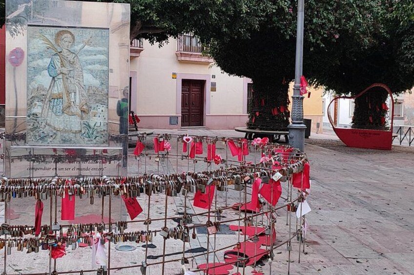 Una plaza pública con un mural grande, una cerca de metal con candados y una cesta roja. 2