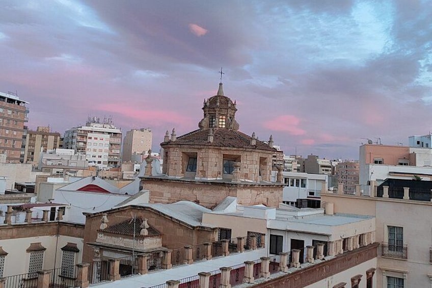 Un edificio histórico con un campanario, rodeado de edificios modernos bajo un cielo rosado. 1