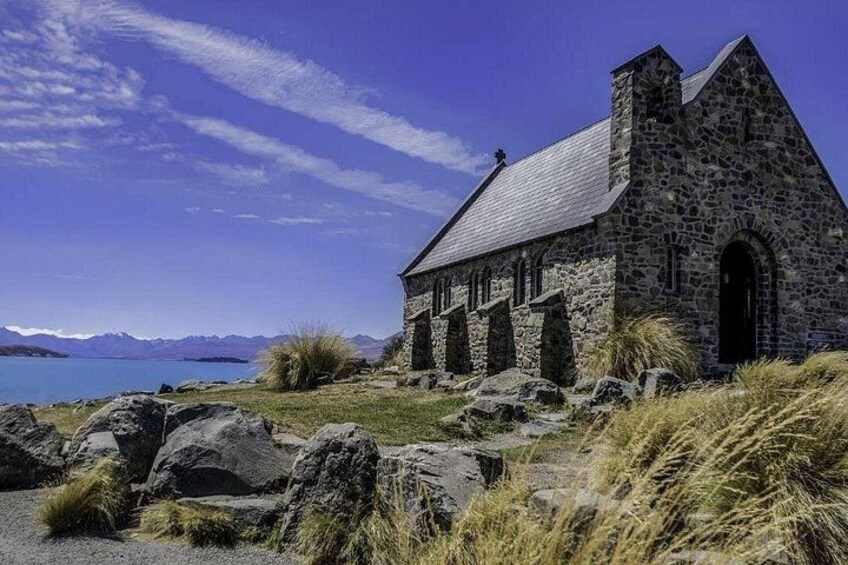 A stone church with a steep roof, situated by a lake with mountains in the background. 5