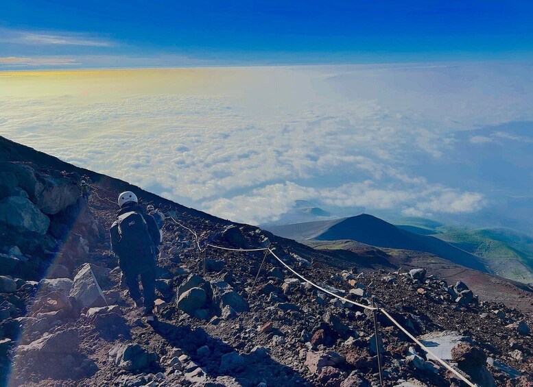 A hiker on a rocky mountain trail with a view of clouds below and distant mountains. 3