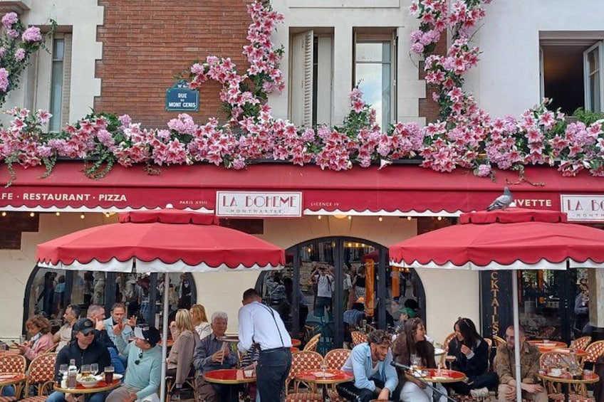 Mesa y sillas al aire libre en un café con toldo rojo y flores rosadas. 5