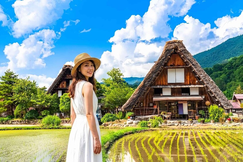 A woman in a white dress and straw hat standing in front of traditional thatched-roof houses with a rice paddy in the foreground. 1