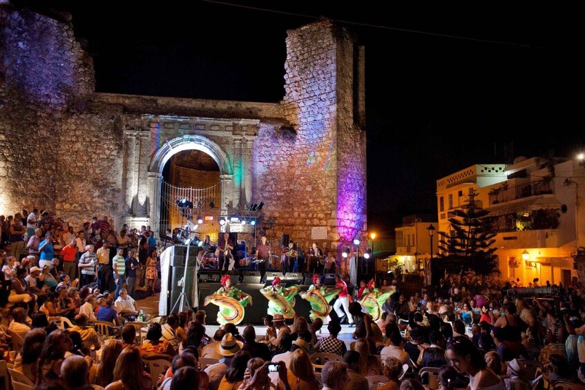 A nighttime outdoor event with a crowd gathered around a stage, featuring performers in colorful costumes. 1