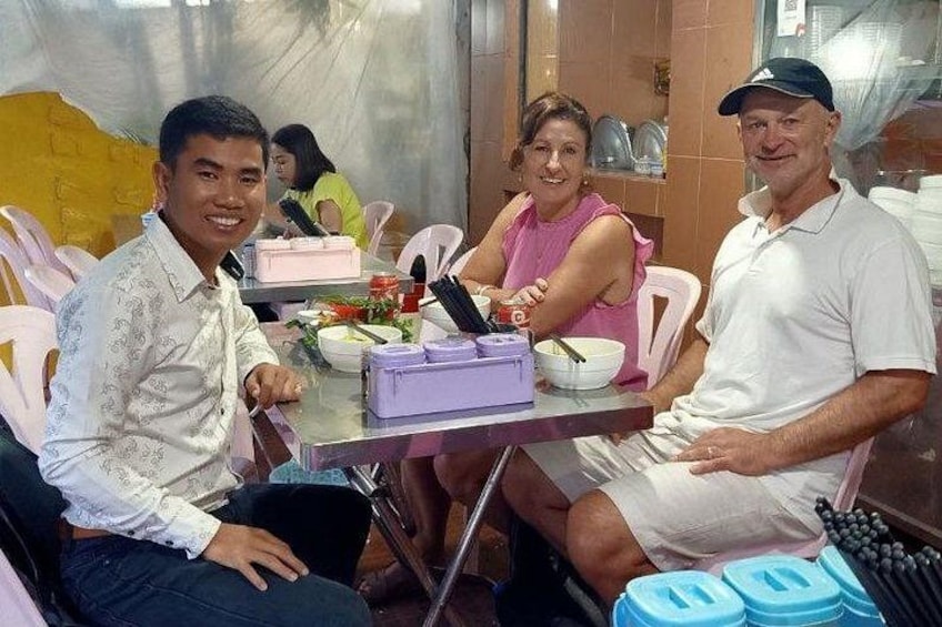 Un grupo de personas sentadas a una mesa con comida y bebidas, en una habitación con pared de azulejos y un objeto amarillo al fondo. 3
