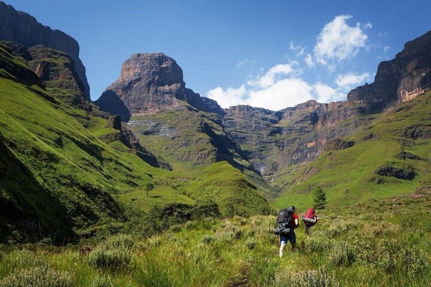 A hiker with a backpack and a red backpack walking on a grassy hillside with mountains in the background. 1