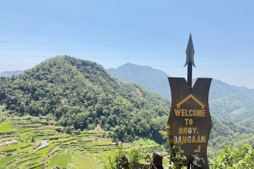 A sign with 'WELCOME TO BRGY. BANGGAAN' against a backdrop of terraced fields and mountains. 3